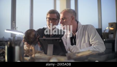 Two archaeologists examine fossil skull, conduct historical and scientific research on remains of ancient extinct human, use digital tablet computer. Scientists work in archaeological laboratory. Stock Photo