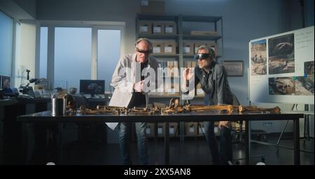 Two archaeologists in VR headsets study remains of ancient extinct human skeleton using augmented reality technology. Scientists doing historical research while working in modern archaeological lab. Stock Photo