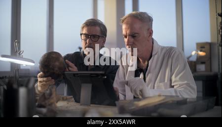 Two archaeologists examine fossil skull, conduct historical and scientific research on remains of ancient extinct human, use digital tablet computer. Scientists work in archaeological laboratory. Stock Photo