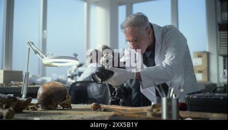 Senior archaeologist takes photo of fossil remains of prehistoric human skull and bones with camera in modern archaeological lab. Scientists work in the background. Archaeology and paleoanthropology. Stock Photo