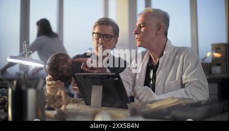 Two archaeologists examine fossil skull, conduct historical and scientific research on remains of ancient extinct human, use digital tablet computer. Scientists work in archaeological laboratory. Stock Photo