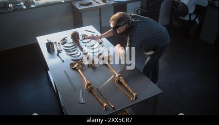 Archaeological laboratory: High angle of mature archaeologist in AR headset studying remains of prehistoric human skeleton. Scientist conducts historical research using virtual reality technology. Stock Photo
