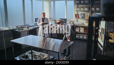 Two archaeologists conduct historical and scientific research in archaeological laboratory using virtual reality technology. Scientist in lab coat uses tablet computer, colleague wears AR headset. Stock Photo