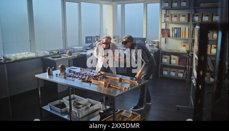 Two archaeologists conduct historical and scientific research in archaeological laboratory using virtual reality technology. Scientist in lab coat uses tablet computer, colleague wears AR headset. Stock Photo