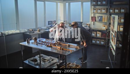 Two archaeologists conduct historical and scientific research in archaeological laboratory using virtual reality technology. Scientist in lab coat uses tablet computer, colleague wears AR headset. Stock Photo