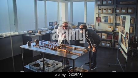 Two archaeologists conduct historical and scientific research in archaeological laboratory using virtual reality technology. Scientist in lab coat uses tablet computer, colleague wears AR headset. Stock Photo