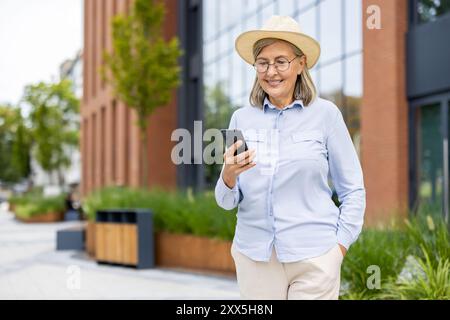 Joyful senior woman wearing hat using smartphone while strolling in city. Modern architecture and lush greenery surround her, reflecting balance between technology and nature. Stock Photo