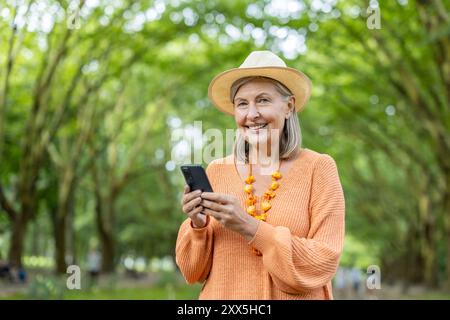 Smiling senior woman using smartphone in green forest. Wearing bright orange sweater and straw hat. Represents joy, technology, and leisure in nature. Embracing new technology and outdoor lifestyle. Stock Photo