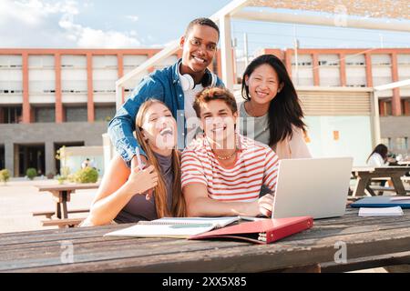 Diverse group of male and female students, smiling and looking at camera while studying together using a laptop on a university campus. Multiracial classmates working on their homework gazing front. High quality photo Stock Photo