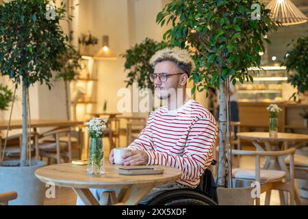 Dreamy reflective disabled man in wheelchair sits with coffee at cafe table. Waits for friend alone Stock Photo