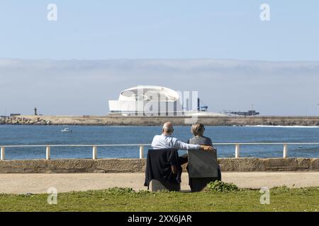 A couple of senior citizens sitting on a park bench looking at the Porto Leixoes Cruise Terminal on the seafront promenade at Praia de Matosinhos bea Stock Photo