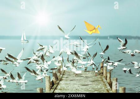 Yellow origami bird and soaring seagulls, jetty on a lake Stock Photo