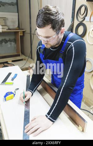 A bearded tired handsome carpenter on a table with a pencil draws a sign on the board. At the workplace there is an ordinary master dressed in blue ca Stock Photo