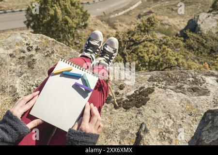 View from the first person on the female lap is a notebook with chalks for drawing pastels. Female hands holding a notebook in nature. A girl sitting Stock Photo