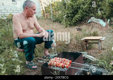 Senior man sitting on chair in garden with grill. Old man on the barbecue on backyard. Senior lifestyle. Meat and tomatoes on skewers on bonfire. Stock Photo