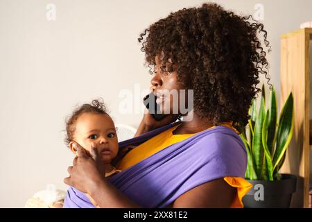 Mother working from home holding baby using sling talking on phone Stock Photo