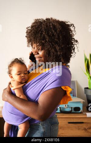 Mother working from home holding baby using sling talking on phone Stock Photo