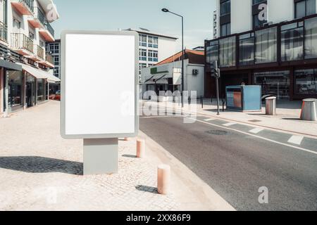 A blank white billboard mockup stands on a deserted suburban street. The poster is positioned on a cobblestone sidewalk with buildings and shops in th Stock Photo