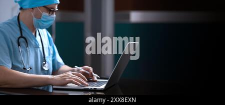 A doctor is sitting at a laptop, working intently. The close-up shot focuses on the laptop and the doctor's hand. Stock Photo