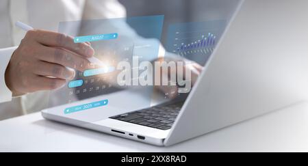 The businessman works at his laptop as a hologram of a calendar appears. Using a stylus, he selects a specific date on the calendar, illustrating his Stock Photo
