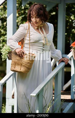A young woman in a white dress and straw hat walks down a garden path, her hand resting on a blue railing. Stock Photo