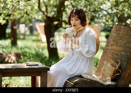 A woman in a white dress enjoys a cup of tea while sitting on a wooden bench in a summer garden. Stock Photo