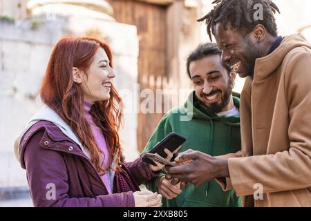 Cheerful multicultural friends looking at smartphone together in urban setting. Young diverse group sharing funny content on social media, representin Stock Photo
