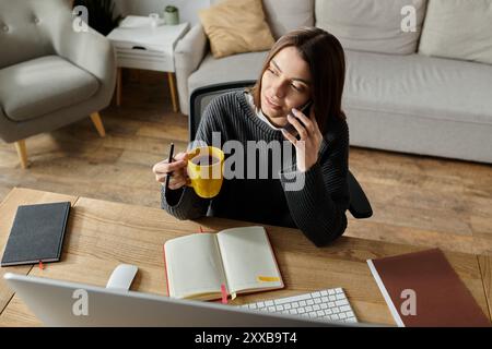 A young woman sits at her desk, working remotely while holding a cup of coffee and talking on her phone. Stock Photo
