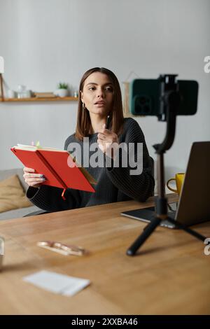 A young woman in a grey sweater sits at her desk, recording a video on her phone while holding a red notebook and looking at the camera. Stock Photo