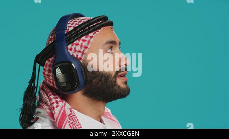 Cheerful guy dancing around in studio, listening to modern tunes on headphones and having fun on camera. Middle eastern adult in traditional clothes showing groovy dance moves and singing. Stock Photo