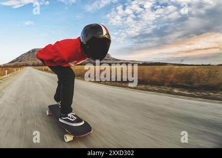A young man in a fullface helmet and a leather suit in a special rack rides a longboard on afsaltu sunset in the background mountains and beautiful sk Stock Photo