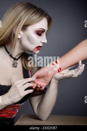 Blonde woman made up as a vampire at Halloween while drinking blood from arm Stock Photo