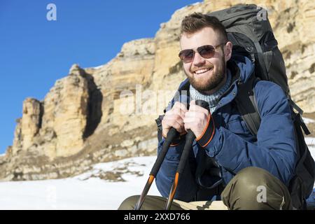 Portrait of happy laughing Hipster traveler with beard in sunglasses is sitting on nature. A man hiking in the mountains with a backpack and Scandinav Stock Photo