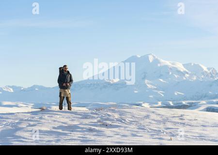 A traveler with a large backpack on his shoulders Stand on a snow-capped hill against the blue sky and the sleeping Elbrus volcano surrounded by the m Stock Photo