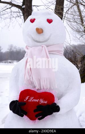 Smiling snowman with heart-shaped eyes holding a red heart with I Love You written on it in snow covered winter scenery. Valentines Day Romantic conce Stock Photo