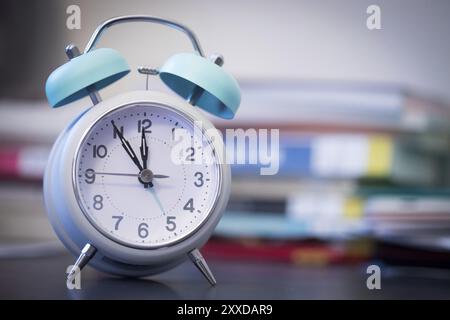 Alarm clock is standing on a grey desk, books and office stuff in the blurry background Stock Photo