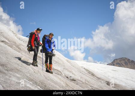 Two tourists, a man and a woman with backpacks and cats on their feet, stand on the ice in the background of the mountains of the sky and clouds. Watc Stock Photo