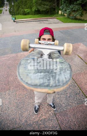 A young long-haired skater in a T-shirt and a full-length cap is standing with a skateboard on the background of the cityscape Stock Photo