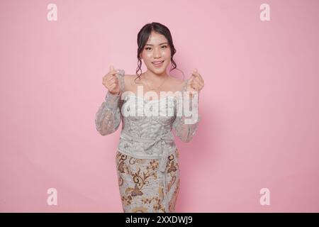 Cute Indonesian woman wearing modern kebaya dress and feeling excited, isolated by a pink background. Stock Photo