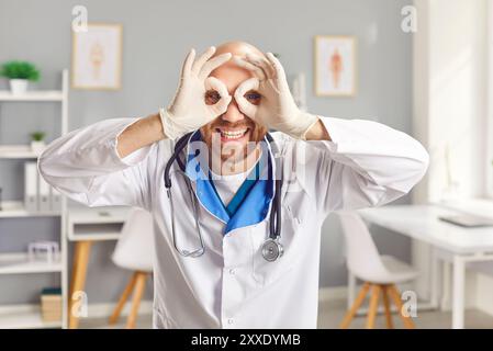 Funny male doctor in modern clinic playfully makes goggles with his hands while smiling widely. Stock Photo