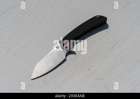 EDC black knife with a red tip sits on a wooden table. The knife is shiny and has a silver blade. The knife is sharp and ready for use Stock Photo