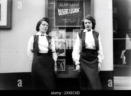 Usherettes standing outside unidentified West German cinema next to coming attraction display for ALAN LADD in DESERT LEGION (1953) at time it was showing DEAN MARTIN and JERRY LEWIS in THREE RING CIRCUS (1954) and LON McCALLISTER and WANDA HENDRIX in MONTANA TERRITORY (1952) in movie theatre showing English language American films for soldiers of US Army of Occupation circa mid-1955 Stock Photo