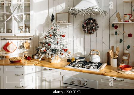 Bright kitchen decorated for Christmas with a frosted mini tree adorned with red and white ornaments, a festive wreath on the wall, and various Stock Photo