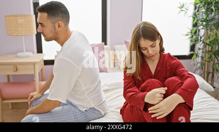 A displeased couple sits apart on a bed within a well-lit bedroom, signifying relationship issues. Stock Photo