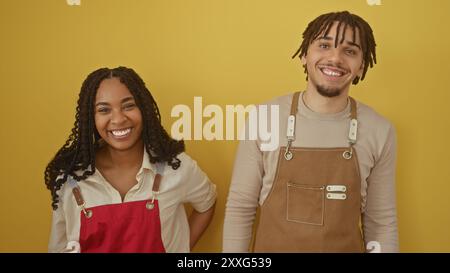A smiling woman and man in aprons standing confidently against a yellow background, depicting a cheerful work environment. Stock Photo