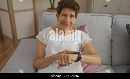 Elderly hispanic woman with short hair sitting indoors on a living room couch, checking her wristwatch with a warm smile. Stock Photo