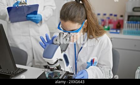 A woman and man work together in a laboratory, with her using a microscope and him taking notes. Stock Photo