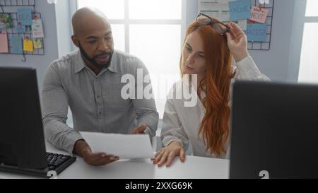 Man and woman collaborating in an office, discussing documents at a desk with computers, highlighting teamwork and business strategy indoors. Stock Photo