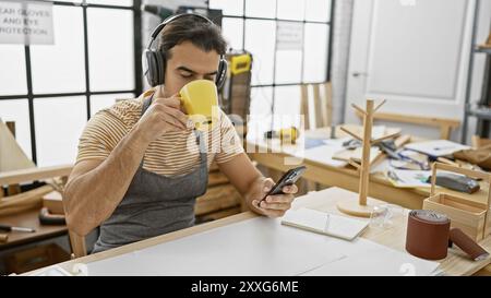 Hispanic man with beard drinking coffee and checking phone in a carpentry workshop Stock Photo