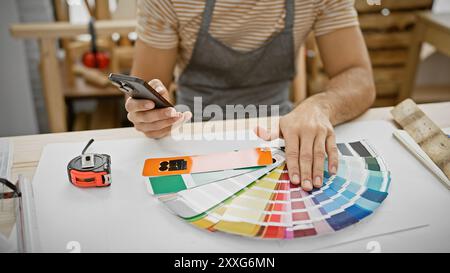A young man examines color swatches in a workshop while using a smartphone, surrounded by carpentry tools. Stock Photo